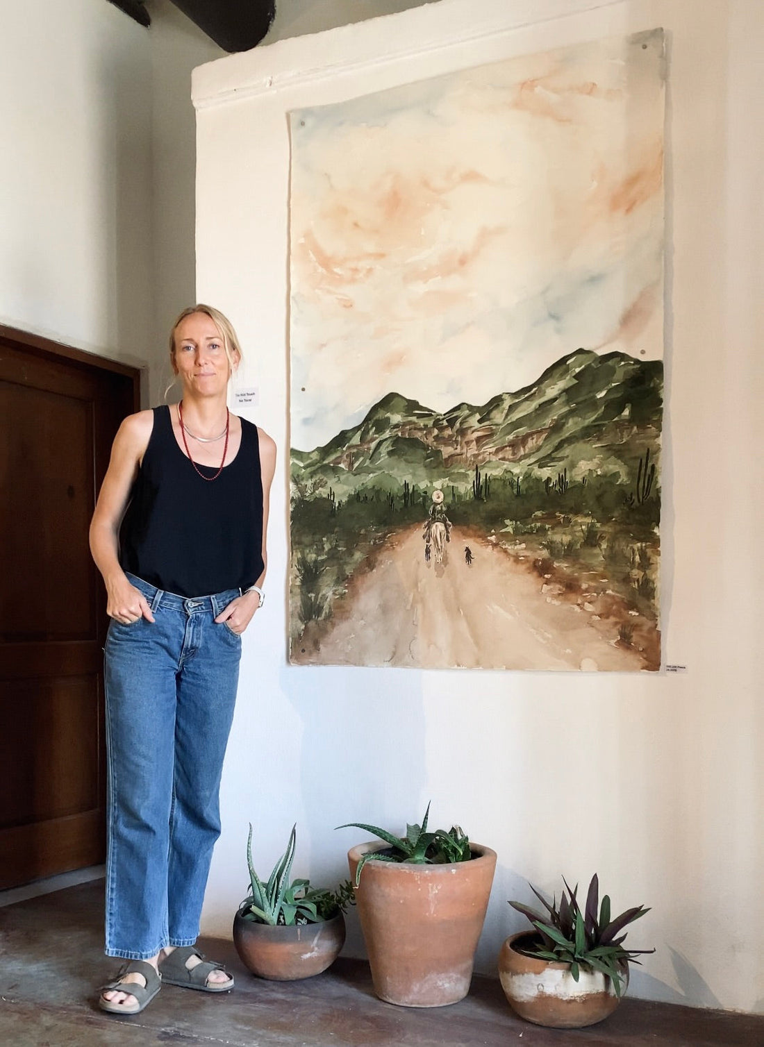 Artist standing beside a large desert landscape painting, with potted plants below, inside an art gallery.